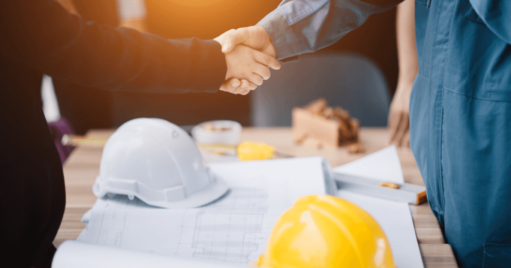 A business woman and construction worker shake hands over a table with a contract to prevent litigation, hardhats, and blueprints